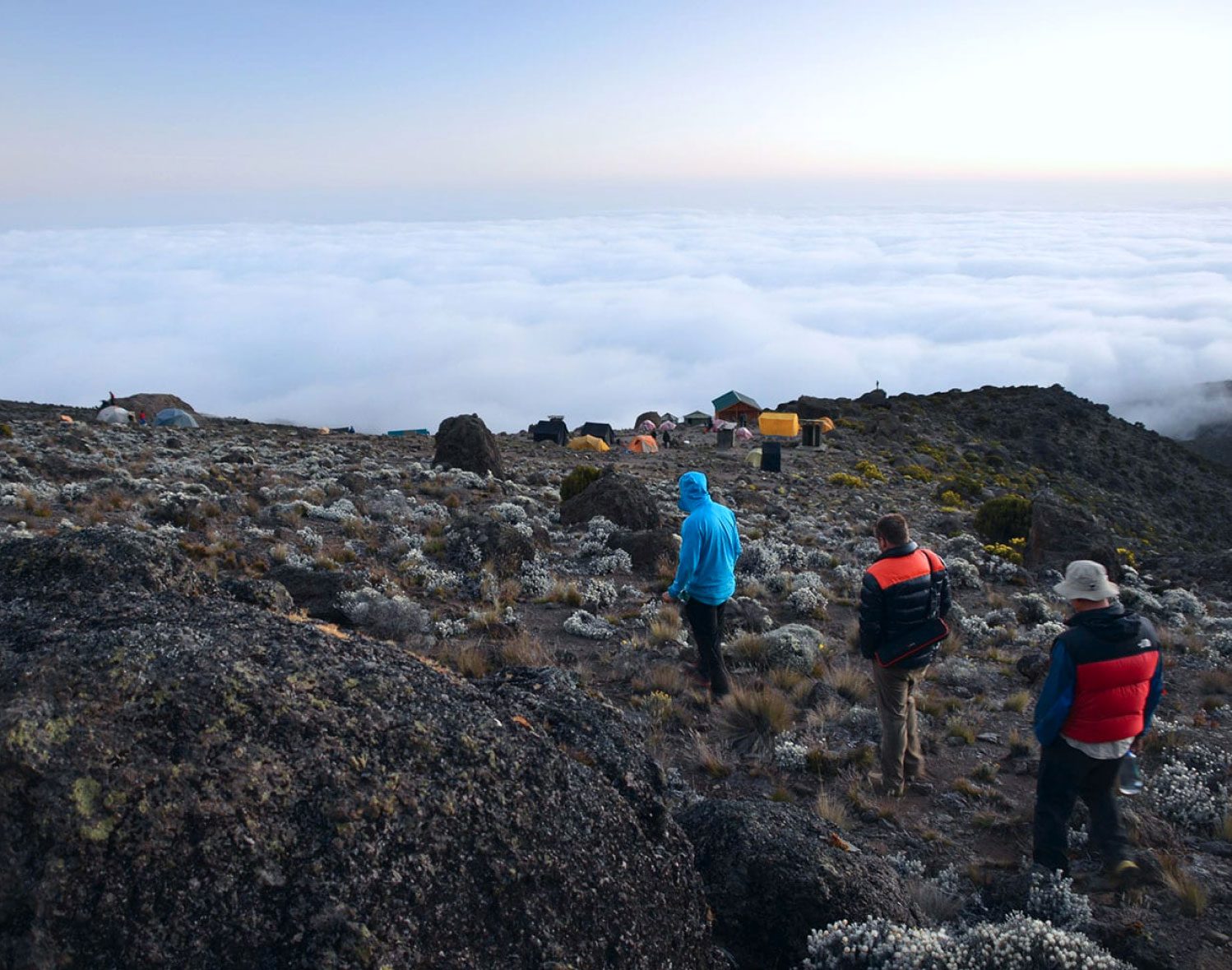 Three young men hike in a line towards a camp while surrounded by clouds on a mountain top.
