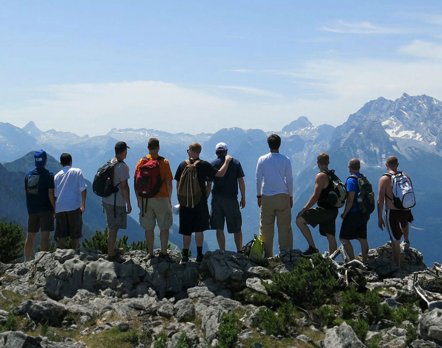 Ten young men look stand in a row across a mountain top and soak in the view of jagged peaks ahead.