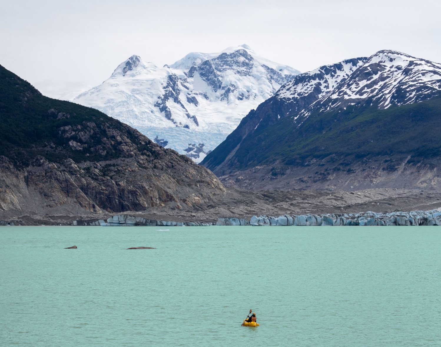 A kayaker paddles across teal colored arctic water in front of a snow covered mountain.