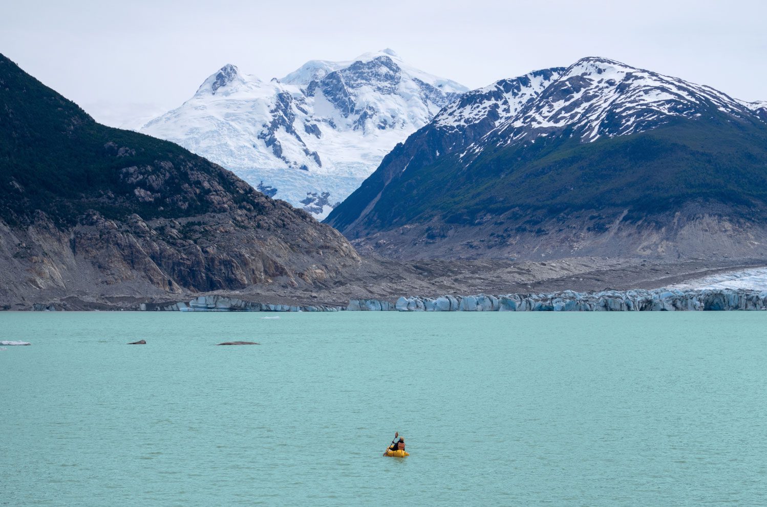 A kayaker paddles across teal colored arctic water in front of a snow covered mountain.