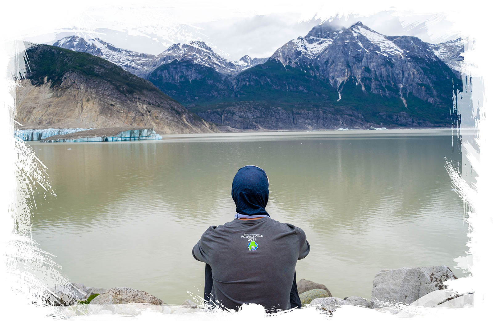 A young man sits alone to think while looking at large mountains over a river in Patagonia, Chile.
