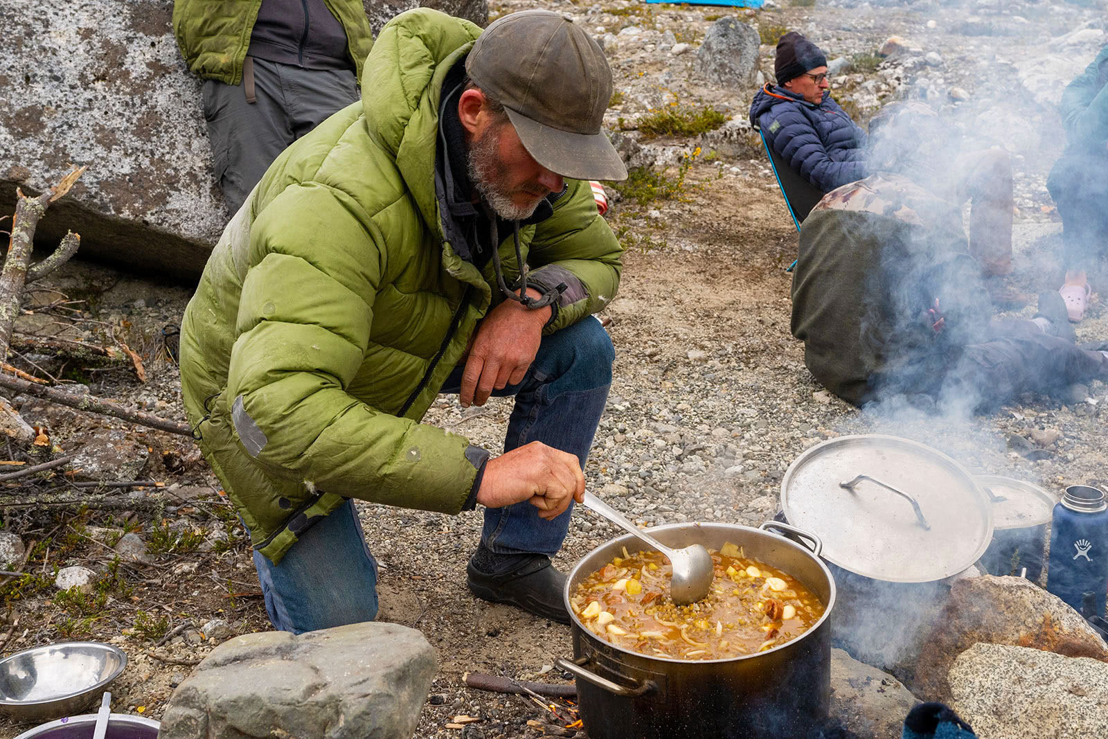 A man in a green jacket cooks a stew over an open fire in Patagonia, Chile.